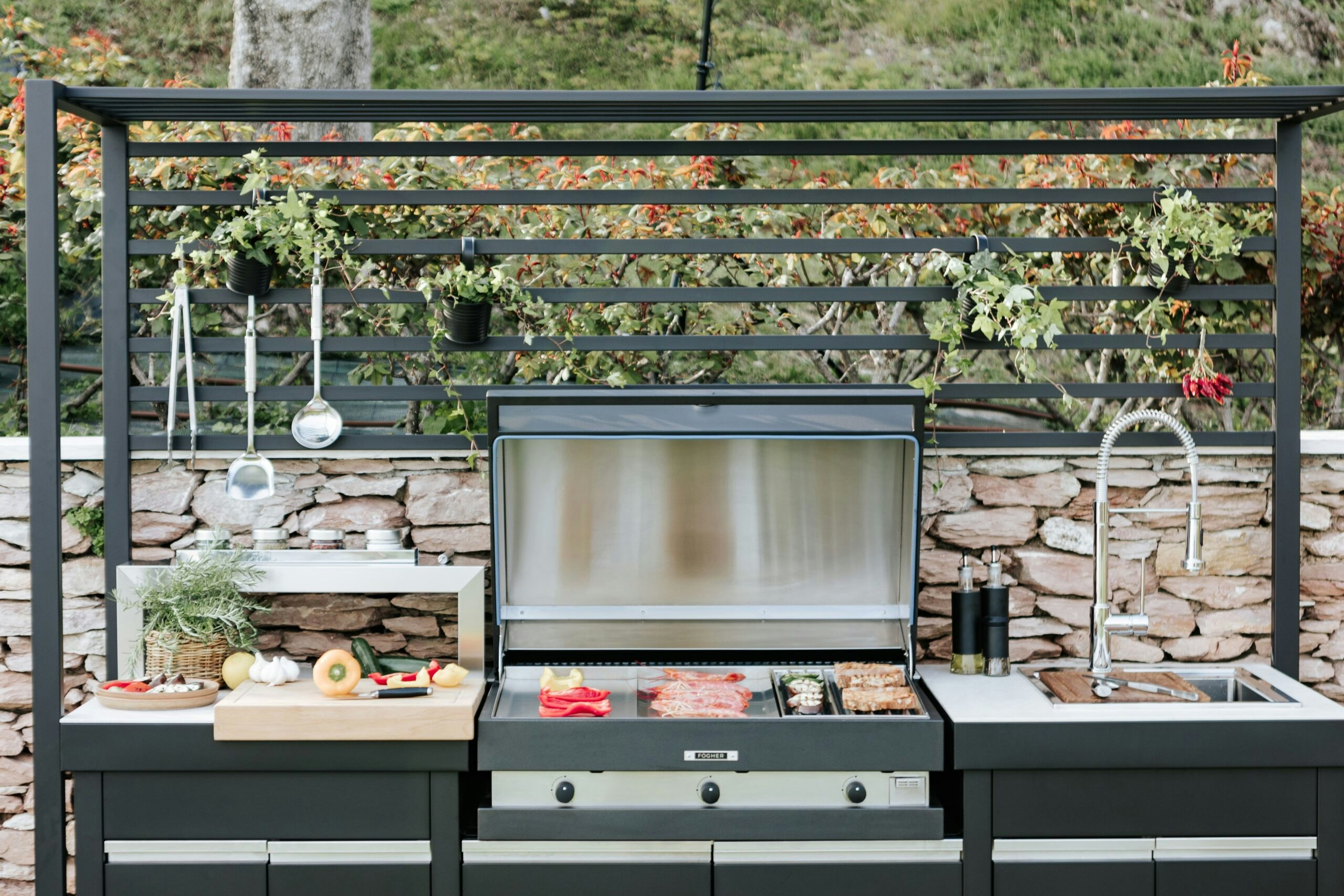 An expansive outdoor kitchen set against a picturesque backdrop. The kitchen area is furnished with modern appliances including a grill, sink, and countertop. The space is adorned by a pergola. Lush greenery and sunlight complete the inviting outdoor culinary space.